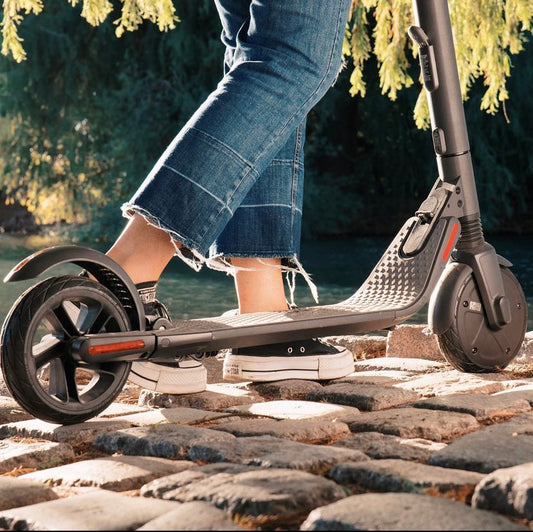 A person stands on a Ninebot electric scooter, resting on cobblestone near a river, showcasing the scooter's deck and wheels.