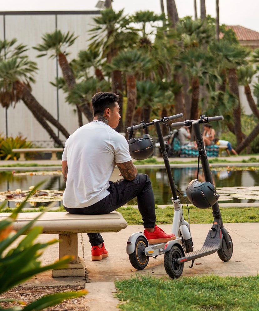 Person sitting on a bench with two Segway electric scooters in a park, emphasizing the convenience and portability of electric scooters.